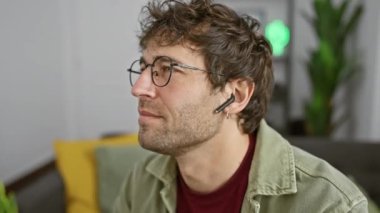 Modern bir oturma odasında gözlüklü, sakallı, kablosuz kulaklık takan İspanyol bir adam..