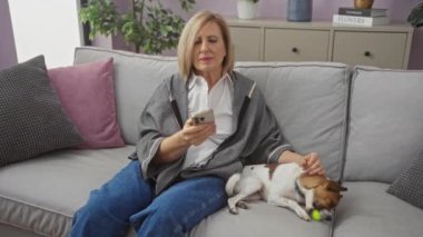 Sarışın, olgun bir kadın oturma odasında oturuyor. Küçük köpeğini gri bir koltukta okşarken akıllı telefonunu kullanıyor..