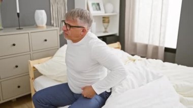Yakışıklı, orta yaşlı, beyaz saçlı, kas ağrısı çeken, ağrıyan sırtına dokunan ve yatak odasında pijama giyen bir adam.