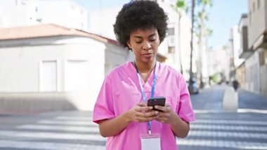 Pembe önlüklü Afro-Amerikalı kadın şehir caddesinde akıllı telefon kullanıyor.