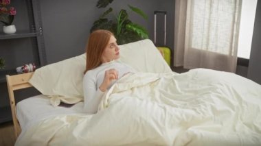 Şaşırmış genç bir kızıl saçlı kadın uyanıyor ve yatak odasında telefonunu kontrol ediyor.