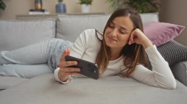 Genç, çekici, sarışın, beyaz bir kadın oturma odasındaki kanepede dinleniyor, rahat kıyafetler giyiyor ve akıllı telefonuna gülümsüyor, rahat ve modern bir ev atmosferi yaratıyor..