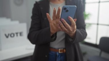 Seçmen odasındaki olgun bir kadın elinde akıllı bir telefonla içeride dikilirken arka planda kısmen belirsiz oy kullanma kabinlerini görüyor..