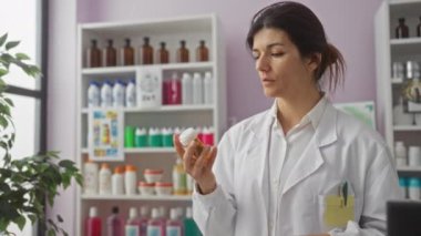 Eczanede beyaz önlük giyen esmer, genç bir kadın eczane içinde iki ilaç şişesini inceliyor..