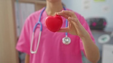 Pembe üniformalı bir kadın hemşire klinik ortamında elinde kırmızı kalp şeklinde bir nesne tutuyor. Tıbbi konuları ve kalp sağlığının önemini vurguluyor..