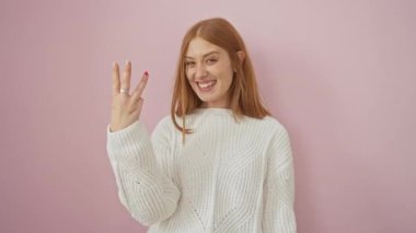 Neşeli genç kızıl saçlı kadın, kendinden emin bir şekilde yukarıyı işaret ediyor, üç numarayı parmaklarıyla gösterirken pozitif ışıldıyor. izole edilmiş pembe bir arkaplan üzerine kaydedildi.