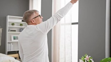 Handsome middle-aged grey-haired man, poised in bedroom, sporting pyjamas, picturesquely pointing towards future whilst posing backwards on bed, a beacon of expectation