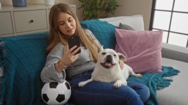 Oturma odasında sarı saçlı genç bir kadın bir telefon tutuyor ve yanında mutlu köpeğiyle bir kanepede oturuyor..