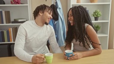 Bir erkek ve bir kadın, sıcak bir evin içindeki ahşap bir masada kahve fincanlarıyla neşeli bir anı paylaşıyor..