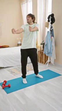 Kafkasyalı sakallı bir adam yoga minderli ve halterli bir odada egzersiz yapıyor.