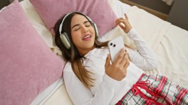 Pijamalı neşeli, genç bir İspanyol kadın yatak odasında kulaklıklarla yatarken akıllı telefonuyla yakınlaşıyor..