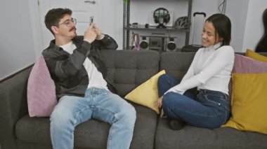 Bir kadın gülerken, bir adam renkli yastıklı rahat bir oturma odasında akıllı telefonuyla fotoğrafını çekiyor..