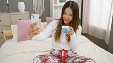 Pijamalı genç bir İspanyol kadın rahat yatak odasında kahve içerken telefonuyla selfie çekiyor..