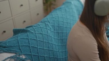 Uzun esmer saçlı genç İspanyol kadın içeride kulaklıklarla müzik dinliyor, rahat bir oturma odasında deniz mavisi bir kanepede dinleniyor..