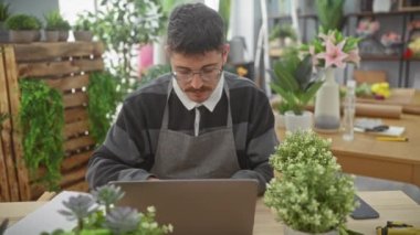 Gözlüklü ve bıyıklı bir İspanyol adam bitkilerle dolu kapalı bir çiçekçide dizüstü bilgisayarda çalışıyor..