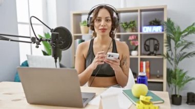 Spor kıyafetli genç bir kadın bitkiler ve dekorlarla dolu modern bir radyo stüdyosunda podcast sunuyor..