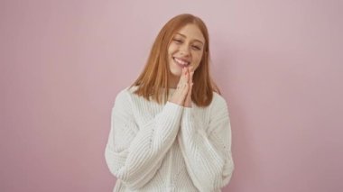 Kendine güvenen genç kızıl saçlı kadın af diliyor, dua ederken ellerini birleştiriyor, pembe izole bir arkaplana gülümsüyor..