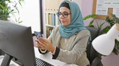 Ofis masasında bilgisayar ve bitki dekoruyla akıllı telefon kullanan tesettürlü profesyonel bir kadın.