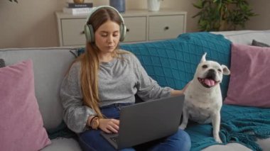Kulaklıklı, sıradan giyinen genç, sarışın bir kadın oturma odasında dizüstü bilgisayarıyla kanepenin üzerinde mutlu köpeğiyle çalışıyor..