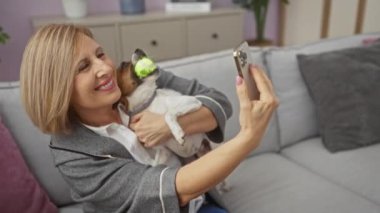 Olgun bir kadın, küçük köpeğiyle sıcak bir oturma odasında selfie çekiyor ve neşeli anlarını yakalıyor.