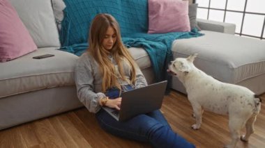 Evde dizüstü bilgisayarı ve akıllı telefonuyla rahatlayan genç sarışın bir kadın. Yanında köpek köpeğiyle rahat bir oturma odasında..