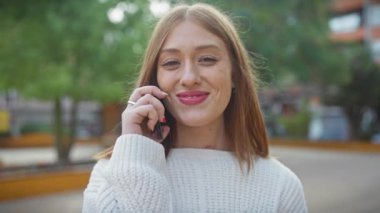 Parktaki bir telefon görüşmesinde gülümseyen kadın süveter giyiyor.