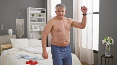 Orta yaşlı bir adam modern bir yatak odasında geriniyor. Yatağında dambıllar var..