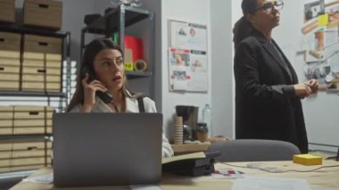 Karmaşık bir soruşturma ofisinde çalışan iki kadın, biri telefonda, diğeri de not alıyor..