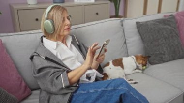Orta yaşlı bir kadın kulaklıkla müzik dinlerken akıllı bir telefon kullanıyor ve rahat bir oturma odasında köpeğiyle kanepede oturuyor..