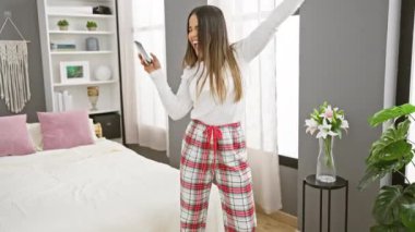 Pijamalı neşeli bir İspanyol kadın şık, modern bir yatak odası ortamında selfie çeker, rahat ve rahat bir ev hayatı tasvir eder..
