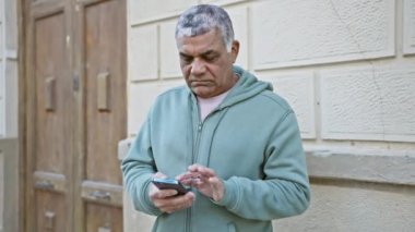 Şehir sokağında arka planda dokulu duvarları olan akıllı telefon kullanan olgun bir adam.