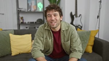 Modern oturma odasında gülümseyen sakallı sıradan İspanyol bir adam, arka planda gitar.