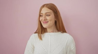 Neşeli genç kızıl saçlı kadın, mutlu bir düşünceye kapılmış, pembe izole bir arka planda duruyor. Bir süveter giyiyor ve tuhaf bir şekilde kendinden emin, ışıl ışıl bir gülümsemeyle yan tarafa bakıyor..