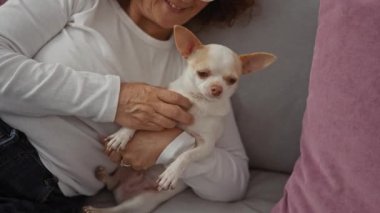 Beyaz gömlekli yaşlı bir kadın oturma odasındaki kanepede pembe yastıklı küçük bir chihuahua köpeğine sarılıyor..