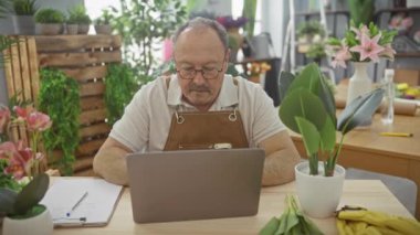 Çiçekçi dükkanında dizüstü bilgisayarda çalışan, etrafı bitkilerle çevrili ve onay veren yetişkin bir adam..