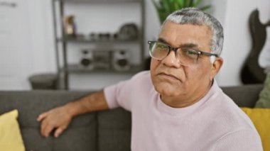 Gri saçlı ve gözlüklü neşeli orta yaşlı bir adam modern bir oturma odasında arka planda gitarıyla gülümsüyor..