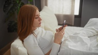 Beyaz kazaklı kızıl saçlı kadın, kapalı bir yatakta otururken akıllı telefonuna çeşitli ifadelerle bakıyor..