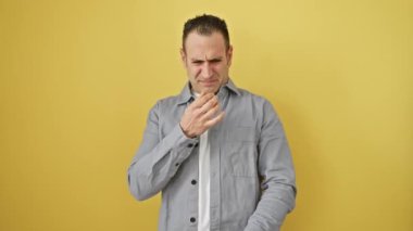 İğrenç kokulu, tişörtlü genç İspanyol adam, dayanılmaz bir kokuyla boğulmuş, nefes tutuyor, burnunu çimdikliyor, saf iğrenme yayıyor, sarı arka planda tek başına duruyor.