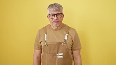 Orta yaşlı, gri saçlı, önlük takan ve gözlüklü bir adam İtalyan jesti yapıyor. El ve parmaklarıyla izole edilmiş sarı arka plan üzerinde kendinden emin bir ifade.