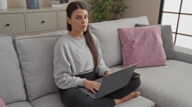 Oturma odasındaki kanepede oturan genç bir İspanyol kadın dizüstü bilgisayar kullanırken parmağıyla el kol hareketi yapıyor..