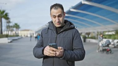 Palmiye ağaçları ve modern mimariyle güneşli bir gezinti alanında akıllı telefon kullanan gülümseyen bir adam.