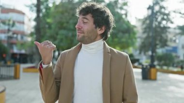 Neşeli genç İspanyol adam, neşeli bir şekilde başparmağıyla yanlamasına işaret ediyor, kendine güveni tam bir şekilde parktaki olumlu duyguları gösteriyor.