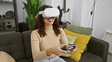 VR kulaklık takan ve elinde kumandayla koltukta oturan gülümseyen kadın.
