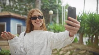 Güneş gözlüklü genç bir kadın arka planda yeşillikle dışarıda selfie çekiyor, günlük ve bağlı bir yaşam tarzını yansıtıyor..