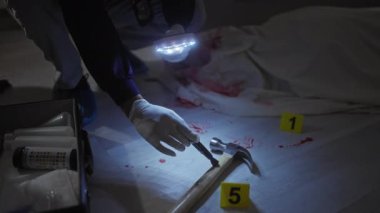 Adamın biri olay yerinde kanlı bir çekiç inceliyor. Kapalı bir yerde, elinde büyüteçle. Soruşturma prosedürlerini gösteriyor..