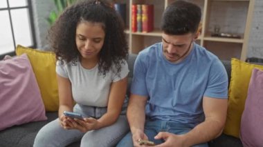 Bir adam ve bir kadın kanepede oturuyor, rahat bir oturma odasında akıllı telefonlar tutuyorlar.