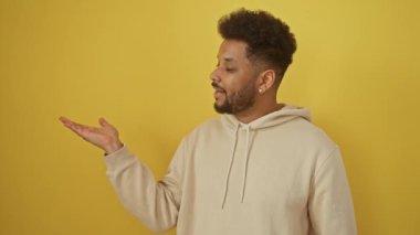 Sarı kapüşonlu neşeli Afro-Amerikalı adam, gülümsüyor ve işaret eden bir el hareketi yapıyor, izole edilmiş sarı arka plandaki kameraya bakıyor.