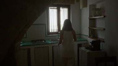 Tipik bir İtalyan evinde genç bir İspanyol kadın, geleneksel bir mutfakta yeşil kiremitler ve beyaz perdelerle duruyor..
