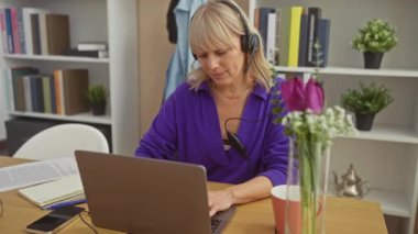 Ev ofisinde dizüstü bilgisayarı, kulaklığı ve çiçekleri olan beyaz bir kadın verimlilik ve rahatlık sergiliyor..