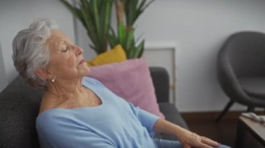 Modern bir oturma odasında renkli yastıklarla rahat bir koltukta dinlenen yaşlı bir kadın..